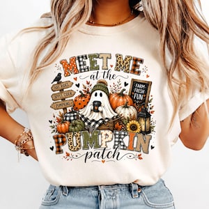 Può includere: Una t-shirt color crema con la grafica di un fantasma che indossa un berretto e tiene in mano una tazza. Il design include zucche, una lanterna, un cartello con scritto Hay Rides, Apple Cider, Pumpkins e il testo Meet Me at the Pumpkin Patch.