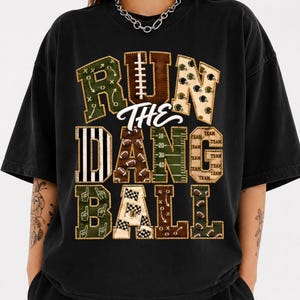 Könnte beinhalten: Ein schwarzes Kurzarm-T-Shirt mit einem großen Grafikdruck und dem Text "RUN THE DANG BALL". Die Buchstaben sind mit verschiedenen Mustern zum Thema Football gefüllt, darunter Spielzüge, Footballs, Helme, Spielfeldmarkierungen und Zielflaggen.