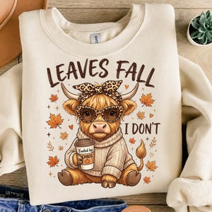 Può includere: Una felpa color crema con la grafica di una mucca Highland che indossa una fascia e occhiali leopardati, mentre tiene una tazza di caffè con la scritta Fueled by CHAOS e una zucca. Il testo recita LEAVES FALL I DON'T.