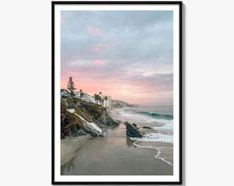 May include: A framed wall art print featuring a coastal landscape at sunset. The scene shows a sandy beach with dark rock formations, gentle ocean waves, and a cliffside topped with white buildings under a soft pink and blue cloudy sky.