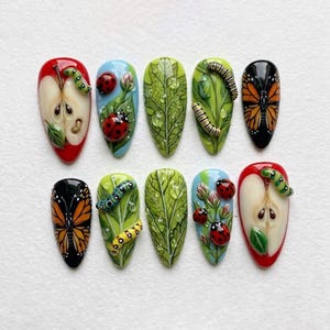 May include: A set of ten artificial nails with detailed, nature-themed designs. The nails feature vibrant colors and 3D elements, including ladybugs, caterpillars, butterflies, and apple and leaf motifs. Each nail showcases a unique miniature artwork.