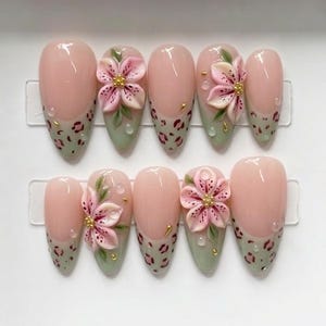 May include: A set of ten almond-shaped press-on nails featuring a nude base with sage green tips. The tips are decorated with a brown leopard print, three-dimensional pink floral accents, small green leaves, and tiny gold metallic studs.