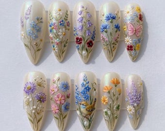 May include: A set of ten almond-shaped artificial nails, each adorned with a unique 3D floral design. The nails feature a pearlescent base with detailed depictions of various flowers in shades of blue, pink, purple, yellow, and white, with a butterfly and bird.