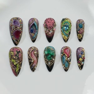 May include: A set of ten almond-shaped, embellished artificial nails. Each nail features a unique design with colorful gemstones, gold-colored metal accents, and intricate details. The nails have a gradient black to purple base.