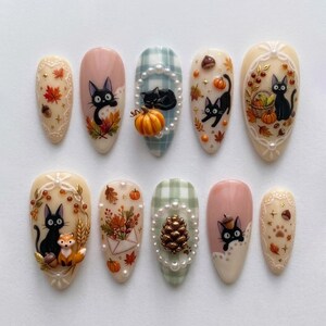 May include: A set of ten almond-shaped press-on nails featuring autumn themes, including black cat illustrations, pumpkins, pinecones, pearl accents, plaid patterns, and fall foliage motifs in shades of orange, brown, cream, and sage green.