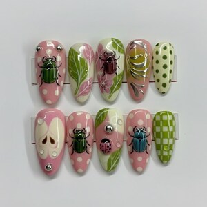 May include: A set of ten artificial nails with various designs. The nails feature pink and white bases with polka dots, green leaves, and insect illustrations. Some nails have silver embellishments.