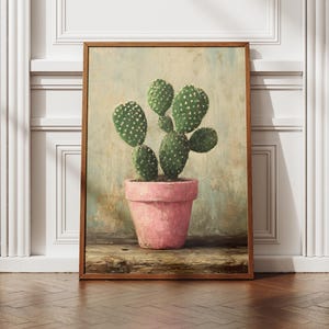 May include: A framed art print featuring a cactus in a pink pot. The cactus has green, rounded pads with white dots. The pot sits on a wooden surface, and the background has a muted, textured appearance. The frame is a warm brown color.