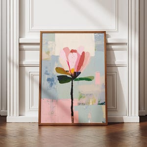 May include: Abstract floral art print in a wooden frame. The artwork features a large pink flower with a black stem and green leaves against a backdrop of pastel colors. The framed art is displayed on a wooden floor.
