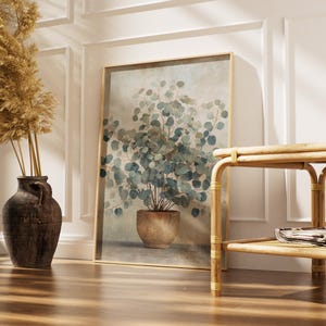 May include: A framed botanical print featuring a watercolor-style eucalyptus plant in a terracotta pot. A dark brown vase with dried pampas grass stands to the left. A light brown rattan side table with a lower shelf is on the right.