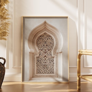 May include: Framed print of an arched architectural detail with intricate, geometric patterns. The artwork features a beige and cream color palette, with a gold frame. The print is displayed in a room with a vase of dried plants.