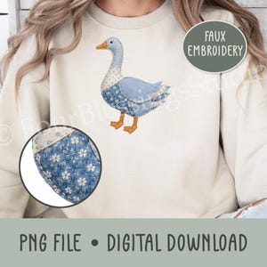 Puede incluir: Una sudadera de cuello redondo color crema con un diseño de bordado falso de un ganso. El ganso tiene el cuello a cuadros azules y blancos, el cuerpo con un estampado floral azul y patas naranjas. Un recuadro circular muestra el patrón floral.