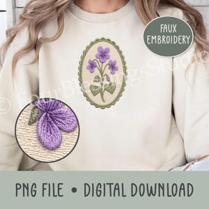 May include: A cream-colored sweatshirt featuring a central oval design with a scalloped green border. Inside the oval is a purple violet flower illustration with green leaves. A circular inset shows a close-up of the purple flower petals.