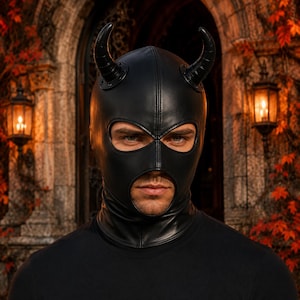 May include: A black leather-like balaclava hood featuring two curved horns attached to the top. The headgear has a large opening for the eyes and a separate circular opening for the mouth, with visible stitched seams running along the center.