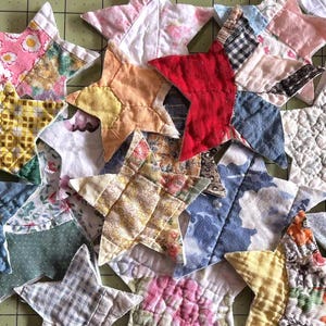 May include: A collection of handmade fabric star ornaments in various colors and patterns. The stars are made from patchwork fabric, featuring floral, geometric, and solid designs. The color palette includes red, yellow, blue, pink, and green.