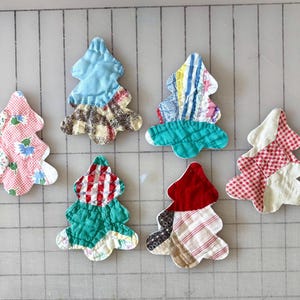 May include: Six quilted fabric ornaments shaped like Christmas trees, each featuring a unique patchwork design. The ornaments display various patterns including red and white gingham, floral prints, solid blue, green, and striped cotton fabrics.
