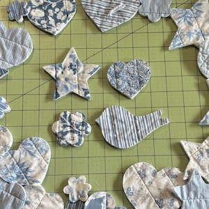 May include: Assorted fabric shapes in blue and white floral and striped patterns. The shapes include hearts, stars, flowers, and a bird, all arranged on a green cutting mat. The fabric has a quilted appearance.