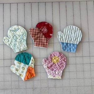 May include: Five small quilted fabric oven mitt ornaments in various patterns and colors. One is light green with a floral print, one is red plaid, one has grey stripes and blue cuffs, one is teal and orange, and one is a pink flower shape.