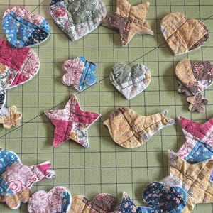 May include: Assorted fabric shapes, including hearts, stars, and a bird, are arranged on a green grid surface. The shapes are made from patchwork fabric in various floral and patterned designs, featuring colors like blue, pink, and yellow.