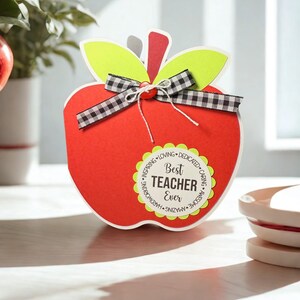 May include: A red apple-shaped gift box with green leaves and a black and white checkered ribbon. The box has a white border and a circular label that reads "Best Teacher Ever."