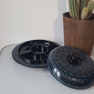 Könnte beinhalten: Schwarze Servierschale mit unterteiltem Innenraum und passendem Deckel. Der Deckel ist mit einem detaillierten, farbenfrohen Muster verziert. Ein Kaktus in einem braunen Topf ist im Hintergrund.