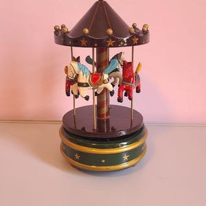 May include: A miniature carousel with three painted horses in red, white, and blue. The carousel has a dark brown roof and base, with gold accents and star details. The base is green with gold trim.