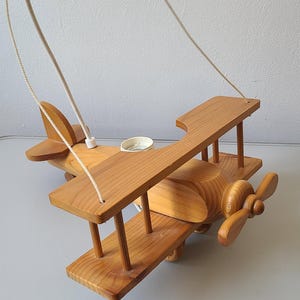 Peut inclure: Un avion jouet en bois avec une hélice, des ailes et une queue. L'avion est en bois de couleur claire et a un design simple. Il est suspendu par un cordon blanc.