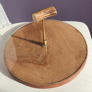 Puede incluir: Una bandeja de servicio redonda de dos niveles con base de madera y tapa de cristal transparente. La bandeja presenta un asa de material texturizado de color claro, posiblemente asta, unida a un vástago de metal color latón. La base de madera tiene un tono marrón cálido.