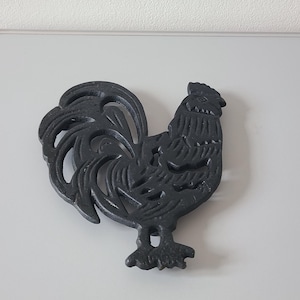 Op de afbeelding: Een zwarte gietijzeren onderzetter in de vorm van een haan. De onderzetter heeft een gedetailleerd ontwerp van een haan met een prominente staart en getextureerde veren. Dit decoratieve item is ontworpen om oppervlakken te beschermen.