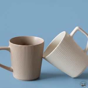 Op de afbeelding: Twee keramische mokken met een verticale geribbelde textuur. De ene mok is gedempt taupe van kleur en de andere is gebroken wit. Beide mokken hebben een handvat aan de zijkant. Een klein rond logo met de tekst Stoneware Pantry Co is zichtbaar.