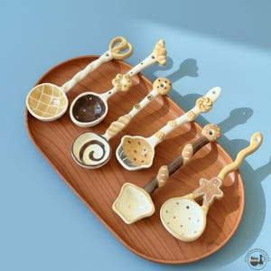 Könnte beinhalten: Fünf dekorative Keramik-Löffel mit verschiedenen Designs, darunter Brezeln und Lebkuchenmännchen, auf einem Holztablett präsentiert. Die Löffel haben cremefarbene, braune und gelbe Farbtöne und gemusterte Schalen. Das Tablett ist etwa 30 cm lang.