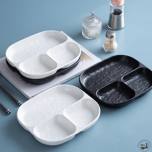 Puede incluir: Tres platos ovalados divididos con superficie texturizada, que incluyen dos platos blancos y uno negro. Cada plato tiene tres secciones distintas para alimentos. Los platos presentan un acabado brillante y un patrón ondulado y orgánico.