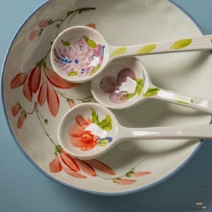 May include: A large ceramic bowl and three matching measuring spoons with hand-painted floral motifs. The bowl features orange and green botanical illustrations, while the spoons display individual pink, purple, and orange flower designs.