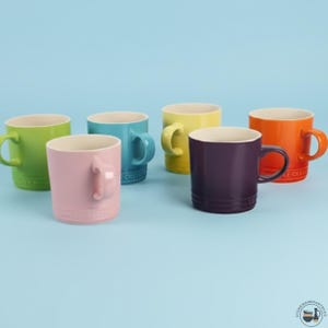 May include: A collection of six ceramic mugs in various colors including lime green, light pink, turquoise, yellow, deep purple, and orange. Each mug features a curved handle, a smooth exterior finish, and a cream-colored interior lining.