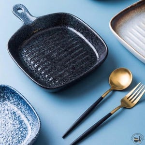 Könnte beinhalten: Ein quadratischer, gesprenkelter schwarzer Keramikservierteller mit Griff und geriffeltem Innenraum. Ein goldener Löffel und eine Gabel mit schwarzen Griffen liegen neben dem Teller. Weitere Keramikgeschirrteile sind im Hintergrund.