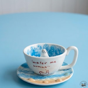 May include: A white ceramic teacup and saucer set featuring a hand-painted blue ocean wave pattern. The cup interior contains a small sculpted white seal figure. The exterior is inscribed with the text "water me human" and a small seal illustration.