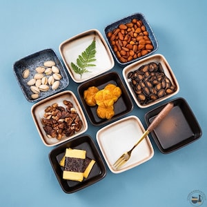Può includere: Un set di otto ciotole quadrate in ceramica in varie tonalità di blu, marrone e crema, disposte su una superficie azzurra. Le ciotole contengono pistacchi, una foglia di felce verde, mandorle, noci, snack gialli, semi neri e una forchetta dorata.
