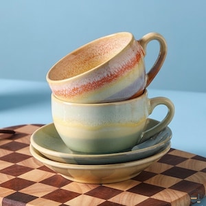 May include: Two stacked ceramic cups with matching saucers. The top cup features a gradient of orange, yellow, and light purple. The bottom cup and saucers display a soft blend of sage green, yellow, and cream, all with a smooth, glossy finish.