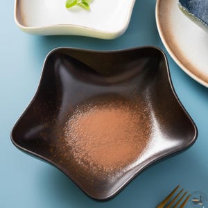 Puede incluir: Un cuenco de cerámica con forma de estrella, exterior marrón oscuro y un centro texturizado con motas de color marrón claro. El cuenco presenta bordes suaves y curvos con un acabado brillante, situado en el centro junto a otros platos de cerámica.