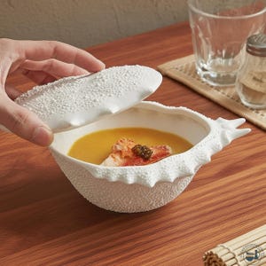 Puede incluir: Cuenco de sopa de cerámica blanca con una superficie texturizada y punteada y una tapa que se levanta. El cuenco contiene sopa naranja, adornada con comida y caviar. El cuenco tiene un diseño único en forma de concha.