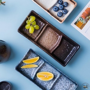 May include: Three rectangular ceramic serving trays with multiple compartments. One dark brown tray holds green grapes, one light beige tray holds blueberries, and one blue speckled tray holds three yellow citrus slices.
