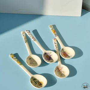 Puede incluir: Seis cucharas de cerámica blanca con diseños florales. Cada cuchara presenta un patrón floral diferente en tonos morados, amarillos y rosas. Las cucharas están dispuestas sobre una superficie azul claro.