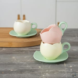 Pode incluir: Três chávenas de chá e pires de cerâmica com um design de tulipa. Uma chávena é branca, outra rosa e a terceira amarelo pálido. Cada chávena tem um pires a condizer e uma pega verde. As chávenas estão empilhadas numa superfície de madeira cinzenta.