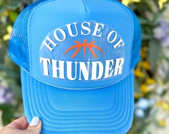 May include: A light blue trucker hat with a mesh back and a curved brim. The hat features the text "HOUSE OF THUNDER" in white block letters with an orange basketball graphic. A hand is holding the hat.