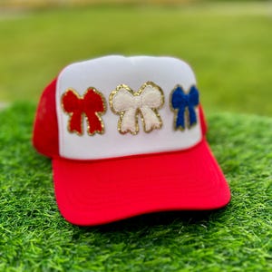 May include: A red and white trucker hat with a red brim and a white front panel. The front panel features three decorative bows in red, white, and blue, each outlined in gold. The hat is likely a patriotic accessory.