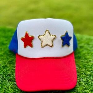 May include: A red, white, and blue trucker hat with three star appliqués. The stars are red, white, and blue with gold trim. The hat has a red brim, a white front panel, and a blue mesh back.