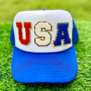 May include: A blue and white trucker hat with the word "USA" in red, white, and blue letters. The letters are outlined in gold glitter. The hat has a blue brim and a white front panel.