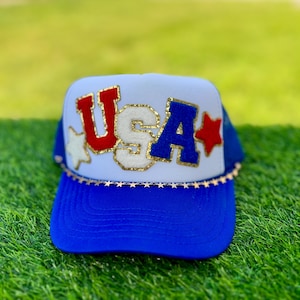 May include: A blue and white trucker hat with the word "USA" in red, white, and blue letters. The letters are outlined in gold glitter. The brim is blue, and the hat is decorated with gold stars.
