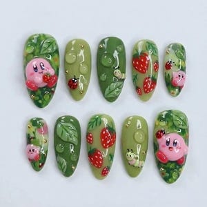 May include: A set of ten artificial nails with a green base, featuring various designs. Some nails showcase a pink character with strawberries, while others display strawberries, ladybugs, and caterpillars. The nails are adorned with leaf and water droplet details.