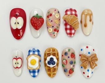 Könnte beinhalten: Zehn künstliche Fingernägel mit verschiedenen Designs zum Thema Essen. Die Designs umfassen einen roten Apfel, eine Erdbeere, Fruchtscheiben, ein Croissant, Sirup, Toast mit Ei, eine Waffel mit Blaubeeren und eine Schleife.