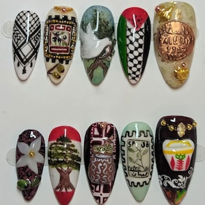 May include: A set of ten artificial nails, each with a unique design. The designs include patterns, flags, and illustrations, featuring colors like white, black, red, green, and gold. Some nails have text and embellishments.